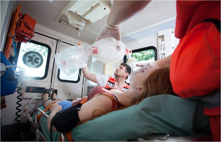 Foto im Krankenwagen, eine Frau liegt auf der Liege am Tropf und Sauerstoffmaske. Ein Sanitäter reguliert den Tropf, der andere am Kopf der liegenden Frau und hält die Sauerstoffmaske fest an ihren Mund. So, war es auch bei mir.